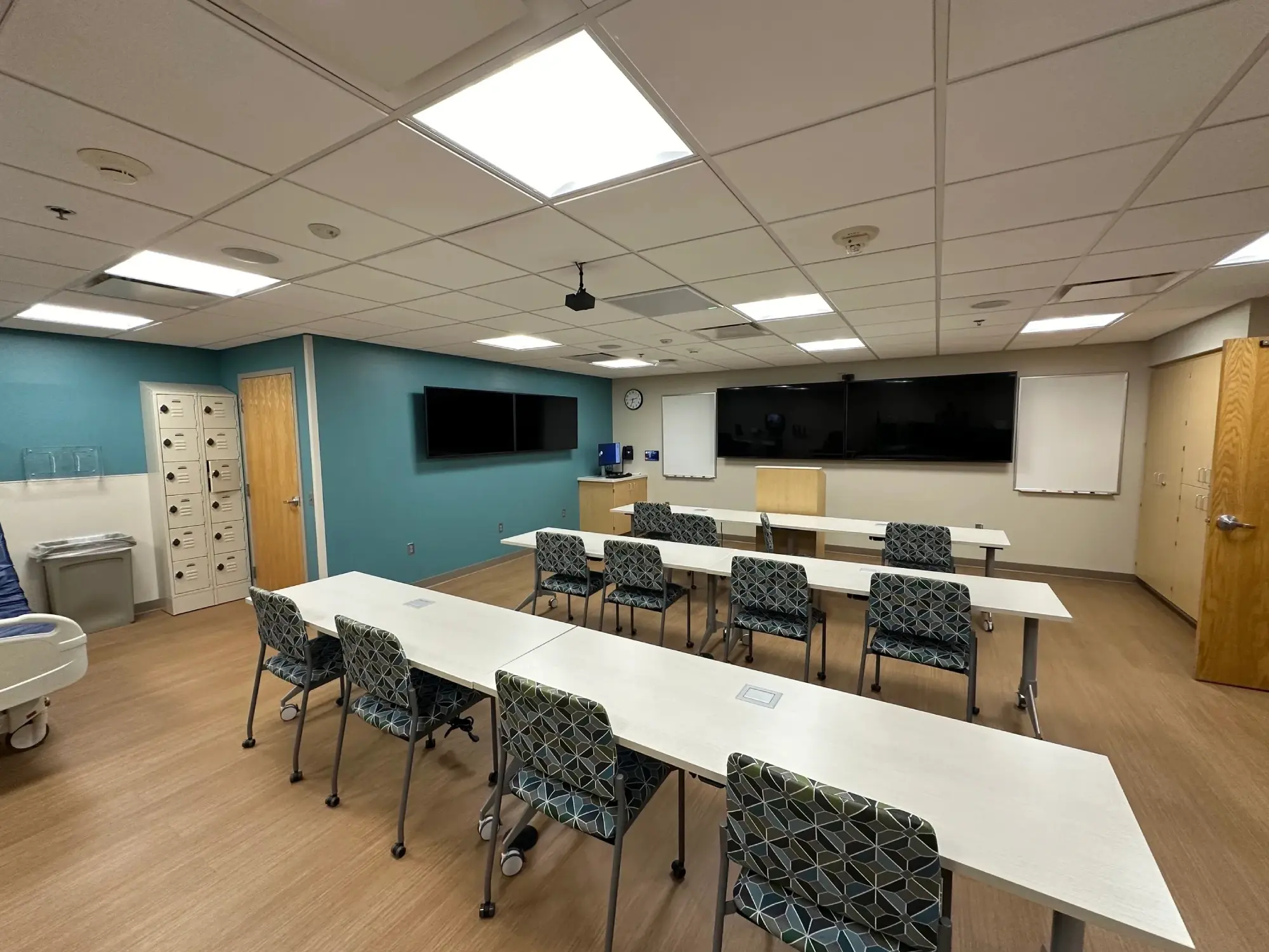 A classroom with three tables and chairs facing two large screens on a wall. The room has a wood floor, lockers, and a sink in the corner.