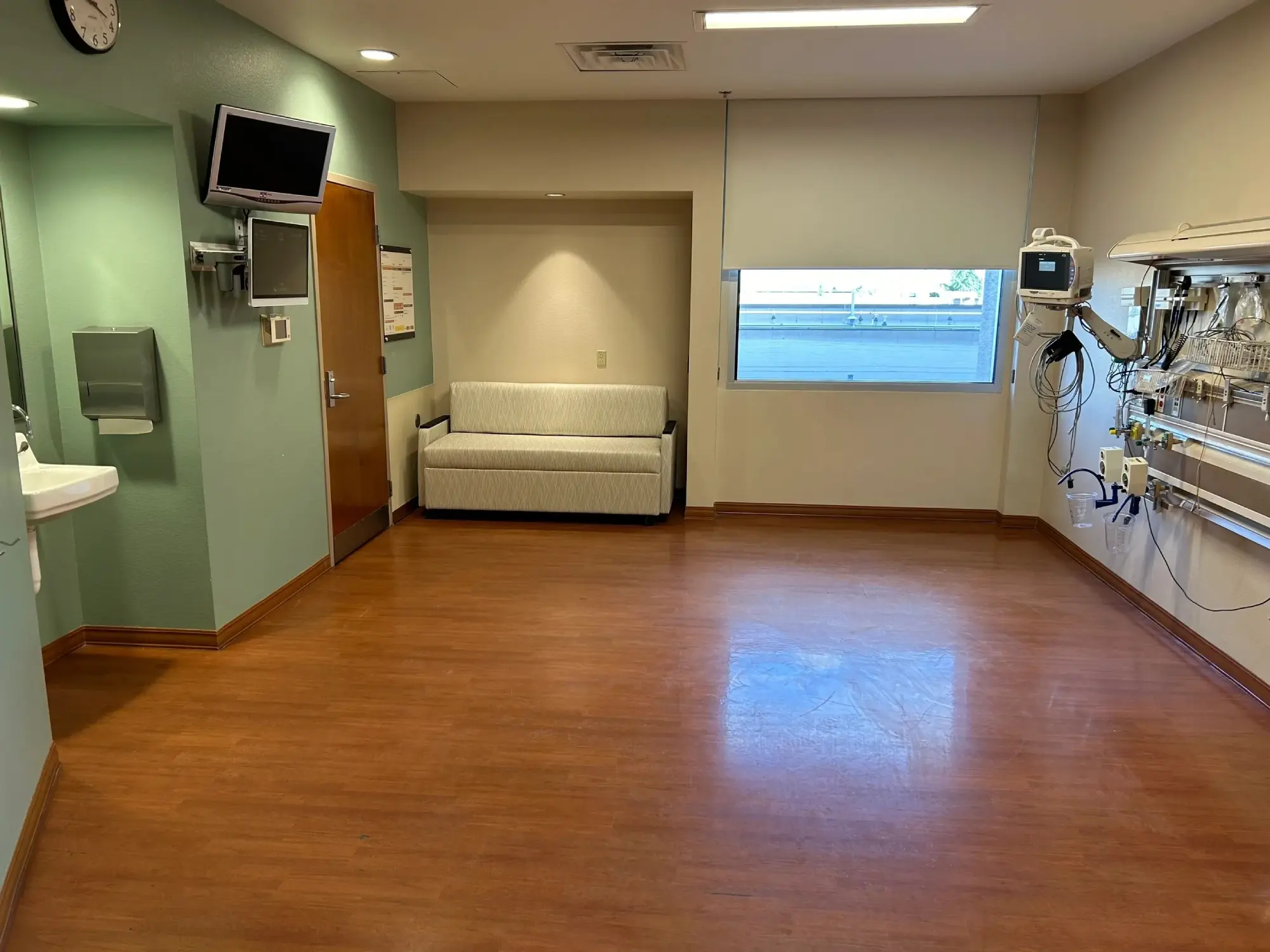 A hospital room with a television, medical equipment, wall clock, sink, and a couch. The room has wooden floors and a window with a blind partially drawn.