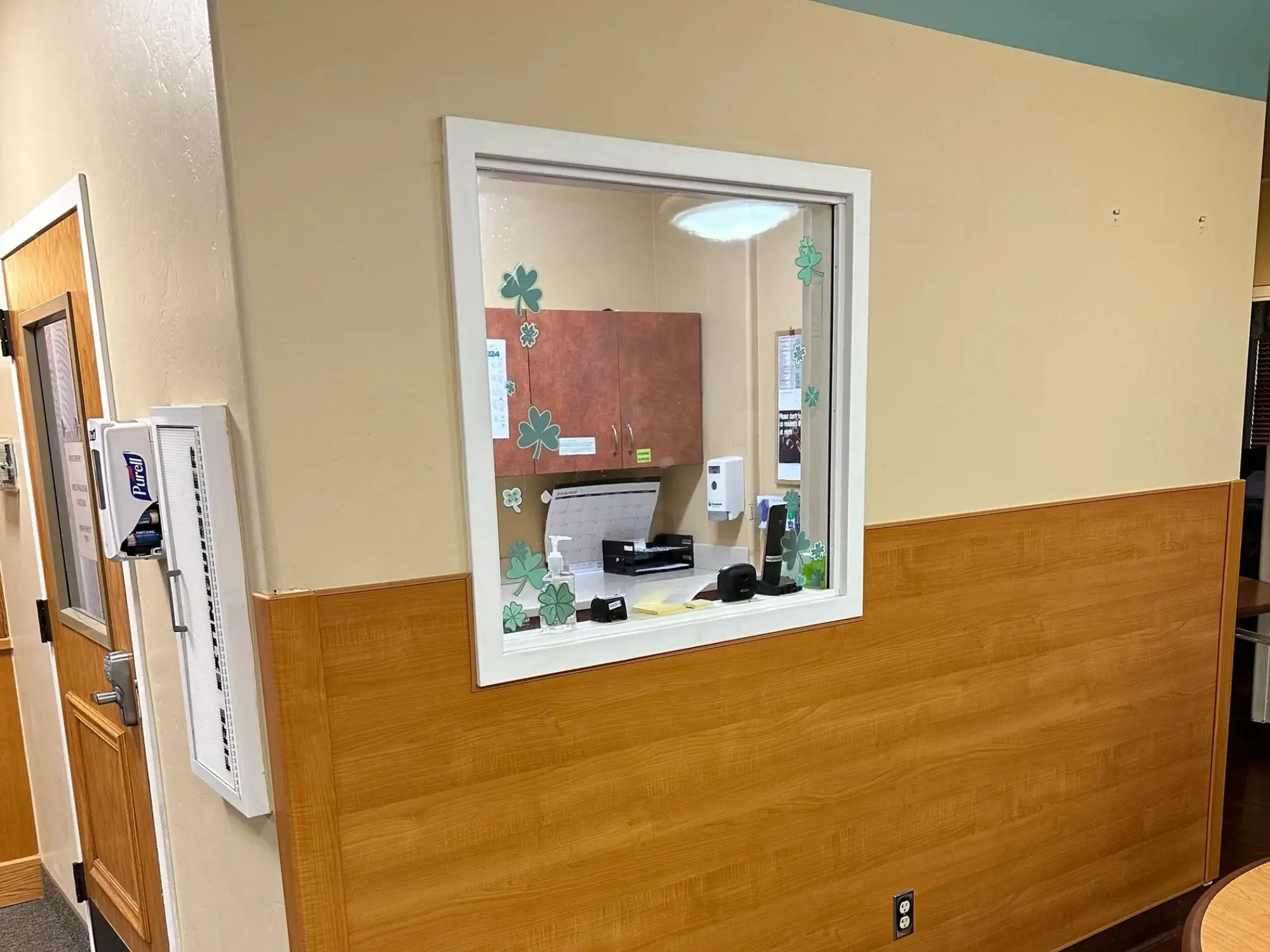 Reception office viewed through a window, decorated with green clover stickers, featuring a desk, computer, and office supplies.