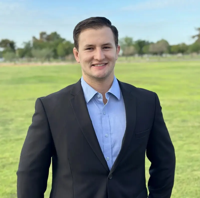 New Project (2) A person in a black suit and light blue shirt stands smiling outdoors with trees and grass in the background.