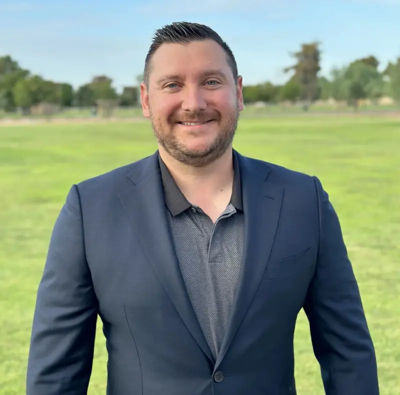 New Project (3) A person in a blue suit and dark shirt stands smiling in a grassy field with trees and a blue sky in the background.