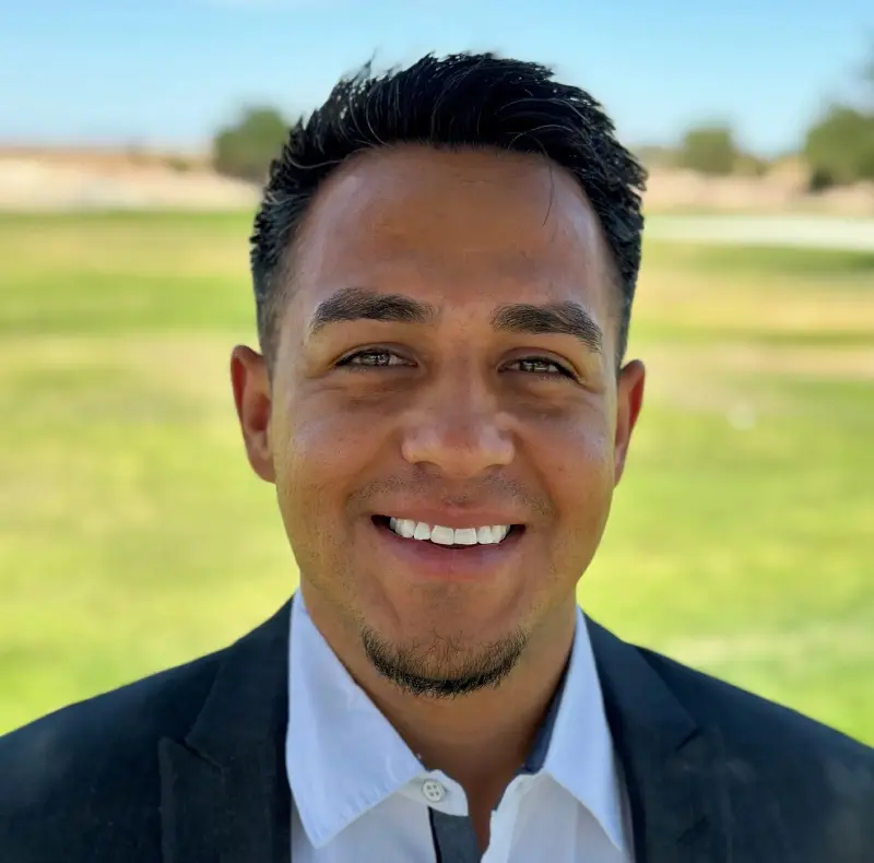 New Project (4) Man smiling outdoors with green grass and trees in the background, wearing a dark blazer and white shirt.